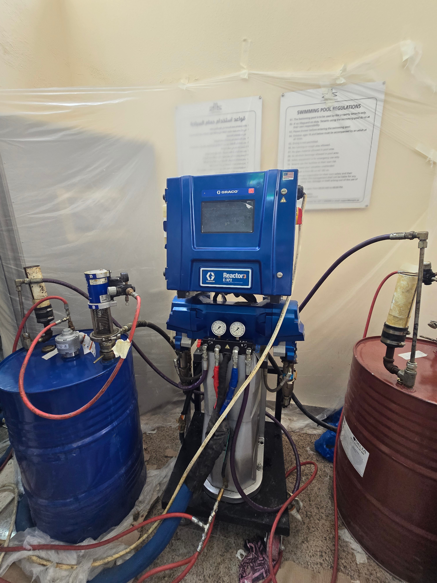 GRACO Reactor 3 E-XP2 plural-component proportioner on site, with Component A (blue drum) and Component B (red drum) feeding heated, high-pressure hoses to the spray gun.